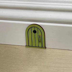 Może przedstawiać: Miniaturowe drzwi do wróżek, pomalowane na jasnozielono z pionowymi liniami i ciemnobrązową, łukowatą ramą. Drzwi mają małe, okrągłe okno i złoty uchwyt. Umieszczone są na białej ścianie i jasnej podłodze.