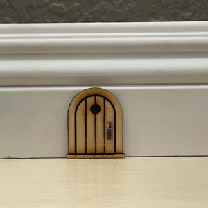 Puede incluir: Una pequeña puerta de hadas de madera con una parte superior arqueada y tablones verticales. La puerta presenta un ojo de buey redondo y un pequeño tirador. La puerta es de color marrón claro y parece estar cortada con láser. Se coloca contra una pared blanca y un suelo de color claro.