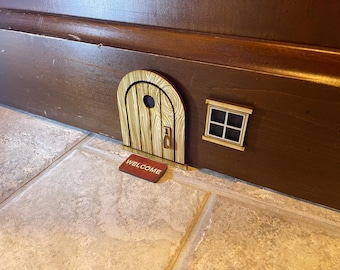 May include: A miniature wooden arched door with a circular window and a small handle, accompanied by a square window with four panes and a rectangular red doormat featuring the word WELCOME in white capital letters, all attached to a baseboard.