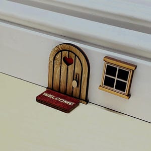 May include: A miniature wooden arched door with a heart-shaped cutout, a round knob, and a rectangular red welcome mat featuring the text WELCOME in white letters, accompanied by a small square window with a dark interior and a wooden frame.