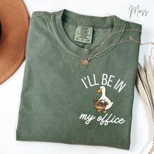 May include: A moss green short-sleeve t-shirt featuring a graphic of a white duck holding a basket of vegetables. The text "I'LL BE IN my office" is printed in white above and below the duck. The shirt has a crew neckline and a tag.