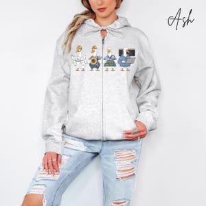 May include: Light grey zip-up hoodie featuring a whimsical design of four geese in various professions. The geese are depicted as a journalist, a construction worker, an electrician, and a computer programmer. The word "Ash" is written in cursive on the upper right.