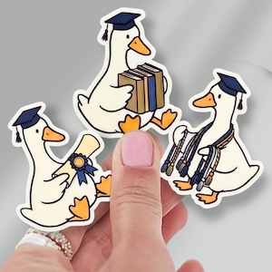 May include: Three die-cut stickers featuring white ducks with orange beaks and feet. Each duck wears a navy blue graduation cap. One duck holds a stack of books, another holds a rolled diploma with a blue ribbon, and the third wears graduation cords.