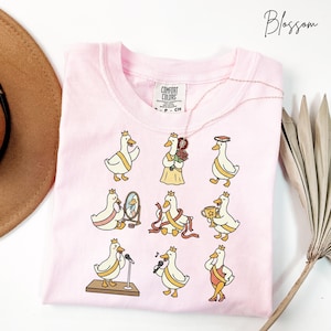 May include: Pink Comfort Colors t-shirt featuring a whimsical design of cartoon geese wearing crowns and various outfits. The geese are depicted in different poses, some holding trophies or microphones. The word "Blossom" is written in cursive.