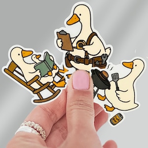 May include: A set of three white duck stickers with cartoon illustrations. One duck reads a book in a rocking chair, another wears a tool belt and holds a clipboard, and the third grills a burger. A can of "Craft Beer" is also visible.