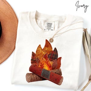 May include: Ivory-colored t-shirt featuring a colorful campfire design. The graphic includes flames in shades of orange, red, and yellow, with logs at the base. A gold necklace with a pendant is draped across the shirt. The shirt has a Comfort Colors tag.