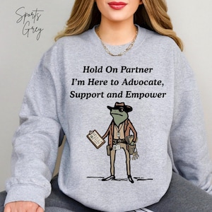 Peut inclure: Sweat-shirt gris Sports avec le texte "Hold On Partner I'm Here to Advocate, Support and Empower." Le sweat-shirt présente une grenouille de dessin animé portant un chapeau, des lunettes de soleil et un costume, tenant un presse-papiers et des clés.
