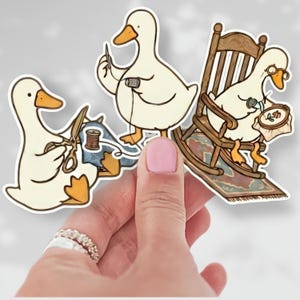 May include: Three die-cut stickers featuring white ducks with orange beaks and feet. One duck uses scissors on blue fabric, one holds a needle and thread, and one sits in a wooden rocking chair while embroidering a floral hoop design.