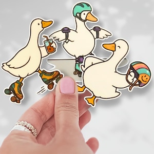 May include: A set of three stickers featuring cartoon ducks on roller skates. One duck holds a drink, another wears a helmet and pads, and the third carries a helmet. The ducks are white with orange beaks and feet, and the roller skates are green and yellow.