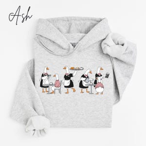 May include: Light gray hoodie featuring a whimsical illustration of geese in a cafe setting. The geese are depicted as waiters and customers, with some holding trays and taking orders. The word "Ash" is written in cursive in the upper left corner.