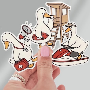 May include: A set of three die-cut stickers featuring white ducks in lifeguard roles. One duck holds a megaphone and a first aid kit, another stands by a wooden lifeguard tower with a life buoy, and the third rides a red and white jet ski.