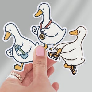 May include: Three white duck stickers with orange beaks and feet. One duck wears a 5k medal, one holds a blue water bottle, and one wears yellow and black running shoes. The stickers feature bold outlines and a cartoon style on a white background.