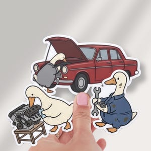 May include: A die-cut sticker featuring three white ducks performing mechanical work. One duck inspects a red car with an open hood, another cleans a car engine on a stand, and the third wears blue coveralls while holding a metal wrench.