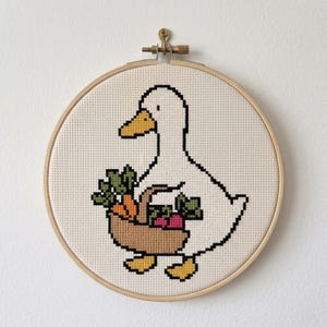 May include: A cross-stitch embroidery of a white duck with a yellow beak and feet, holding a tan woven basket filled with green leafy vegetables, orange carrots, and pink radishes, secured within a round wooden embroidery hoop frame.