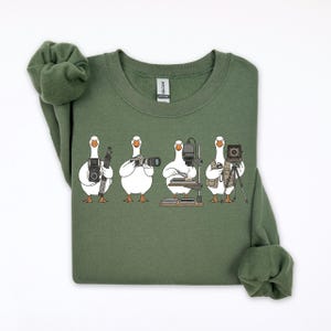 May include: An olive green sweatshirt featuring a whimsical illustration of four white geese, each holding different types of cameras. The geese are depicted as photographers, with various camera equipment. The sweatshirt has a crew neck and long sleeves.