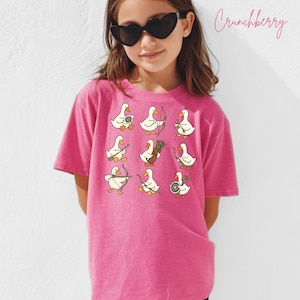 May include: A bright pink t-shirt featuring a pattern of cartoon ducks with bows and arrows. The ducks are in various poses, some holding bows, others with quivers. The shirt has a relaxed fit and is worn by a person wearing heart-shaped sunglasses.