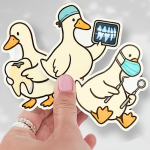 May include: A set of three die-cut stickers featuring cartoon ducks in dental-themed roles. One duck holds a tooth, another an X-ray, and the third wears a mask and holds dental tools. The ducks are cream-colored with orange beaks and feet.