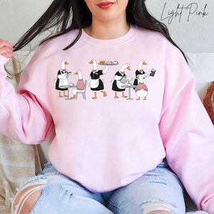 May include: Light pink sweatshirt featuring a whimsical illustration of geese dressed as waiters and customers in a cafe setting. The geese are depicted in various poses, serving food and drinks. The words "Light Pink" are written in cursive in the upper right corner.