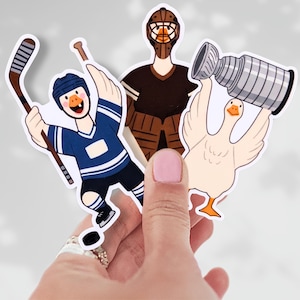 May include: A hand holding three stickers featuring cartoon geese in hockey gear. One goose wears a blue jersey and holds a hockey stick, another is a goalie, and the third goose holds a trophy. The stickers have a white border.