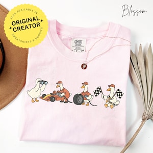 May include: A light pink Comfort Colors t-shirt featuring a cartoon design of racing geese. One goose holds binoculars, another works on a race car, a third changes a tire, and the last waves checkered flags. A gold necklace and a yellow sticker are also visible.
