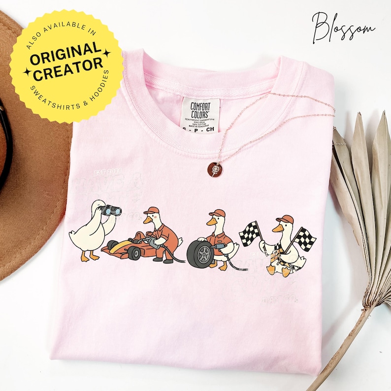 May include: A light pink Comfort Colors t-shirt featuring a cartoon design of racing geese. One goose holds binoculars, another works on a race car, a third changes a tire, and the last waves checkered flags. A gold necklace and a yellow sticker are also visible.