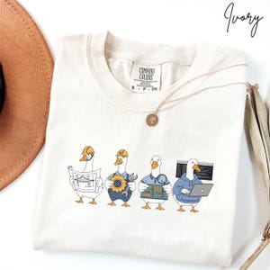 May include: Ivory t-shirt with a graphic of four cartoon geese representing different professions: architect, mechanic, electrician, and programmer. The shirt has a crew neck and short sleeves. A gold necklace is draped across the shirt.