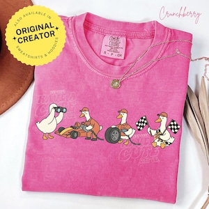 May include: A pink Comfort Colors t-shirt featuring a cartoon of ducks involved in a race. One duck uses binoculars, another works on a race car, another changes a tire, and another waves checkered flags. The text "EST 2024" is visible.
