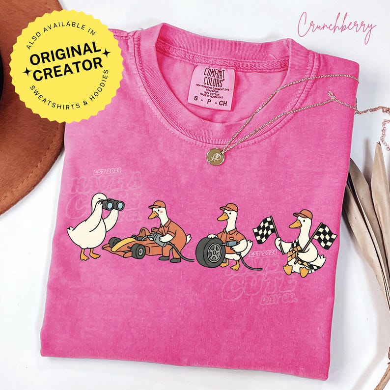 May include: A pink Comfort Colors t-shirt featuring a cartoon of ducks involved in a race. One duck uses binoculars, another works on a race car, another changes a tire, and another waves checkered flags. The text "EST 2024" is visible.