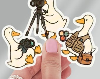 May include: A collection of three stickers featuring cartoon ducks in various photographer roles. One duck holds a camera, another uses a camera on a tripod, and the third wears a vest and carries a bag. The ducks are white with yellow beaks and feet.