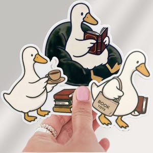 May include: A set of three white duck stickers with orange beaks and feet. One duck sits in a dark green chair reading a book, another holds a steaming cup of coffee, and the third carries a tan tote bag labeled BOOK TOTE and a brown book.