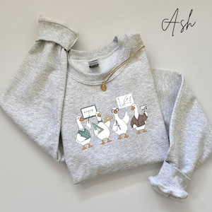May include: Light gray sweatshirt with a graphic of four geese, each holding a sign or wearing a jacket, representing different academic subjects. The geese are wearing glasses and holding a protractor, a sign with the equation x+y=z, and a graph. A gold necklace is draped across the sweatshirt.