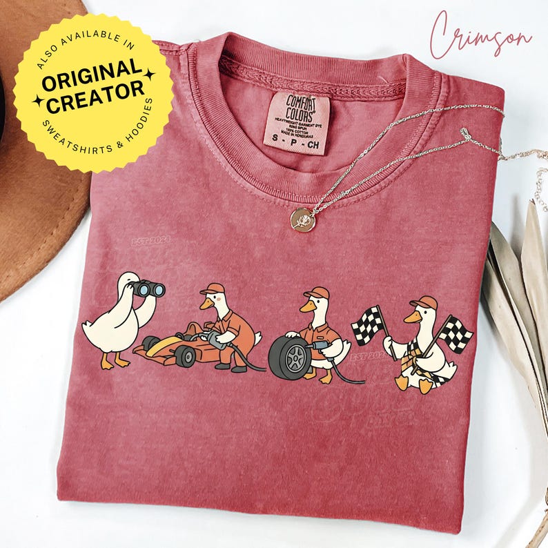 May include: Crimson-colored t-shirt featuring a cartoon of ducks involved in a race. One duck uses binoculars, while others work on a race car, change a tire, and wave checkered flags. The shirt has a Comfort Colors label.