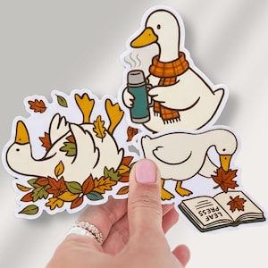 May include: Three die-cut stickers featuring white ducks. One duck wears an orange plaid scarf and holds a teal thermos. Another duck lies in a pile of autumn leaves. The third duck looks at an open book labeled LEAF PRESS with a leaf inside.