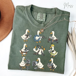 Puede incluir: Camiseta verde musgo Comfort Colors con un diseño caprichoso de patos blancos en varios roles de electricista. Los patos se representan con herramientas y equipos, creando un gráfico lúdico y único.