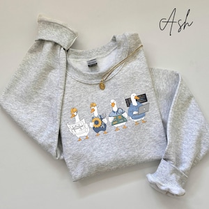 May include: A pale grey sweatshirt with a design of four cartoon geese in different work roles. One goose wears a hard hat and holds blueprints, another holds a wrench, another holds a circuit board, and the last one is on a laptop. A gold necklace is draped across the sweatshirt.