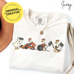 May include: Ivory-colored t-shirt featuring a cartoon design of ducks in racing attire. The ducks are depicted with binoculars, driving a race car, changing a tire, and waving checkered flags. The shirt has the text "Have a Great Race Day".
