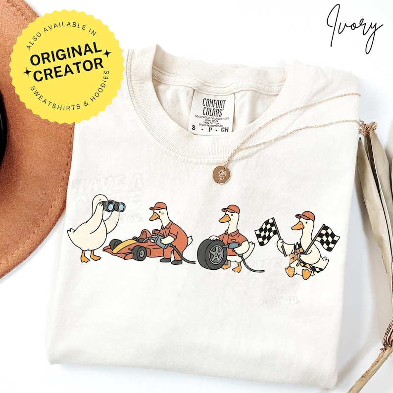 May include: Ivory-colored t-shirt featuring a cartoon design of ducks in racing attire. The ducks are depicted with binoculars, driving a race car, changing a tire, and waving checkered flags. The shirt has the text "Have a Great Race Day".