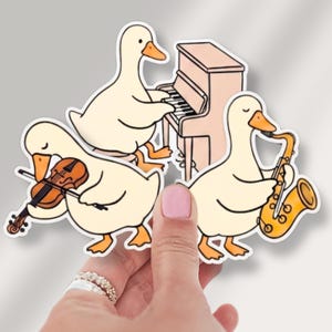 May include: A die-cut sticker featuring three cream-colored ducks playing musical instruments. One duck plays a brown violin, another plays a light pink upright piano, and the third duck plays a bright yellow saxophone. The illustration has a white border.