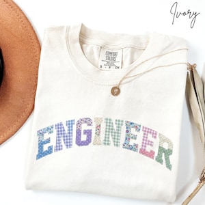 May include: Ivory-colored t-shirt with the word "ENGINEER" in colorful floral and patterned letters. The shirt has a crew neck and short sleeves. A gold necklace with a pendant is draped across the shirt.