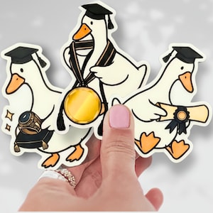 May include: Three stickers featuring cartoon geese celebrating graduation. Each goose wears a black graduation cap. One goose holds a diploma, another a ring on a pillow, and the third wears a medal. The geese are white with orange beaks and feet.