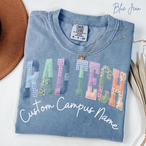 May include: Blue jean colored t-shirt with the words "RAD TECH" in patchwork letters. The letters are in various patterns and colors. Below the text is the phrase "Custom Campus Name" in a script font. A gold necklace is draped across the shirt.