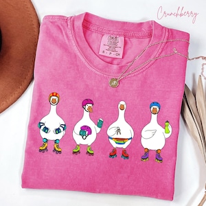May include: Pink Comfort Colors t-shirt with a graphic of four cartoon geese wearing roller skates and helmets. Each goose has a different accessory, including wrist guards, a helmet, keys, and a water bottle. The word "Crunchberry" is written in cursive.