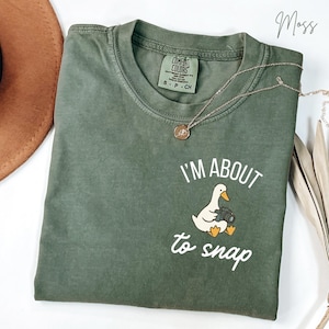 May include: A moss green short-sleeve t-shirt featuring a graphic of a white duck holding a black camera. The text above and below the duck reads I'M ABOUT to snap in white lettering. The shirt has a crew neckline and a Comfort Colors brand label.