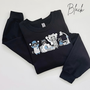 May include: Black sweatshirt featuring a graphic of raccoons in medical attire, including a skeleton raccoon. The design includes medical equipment and the word "Black" in cursive. The sweatshirt has long sleeves and a crew neck.
