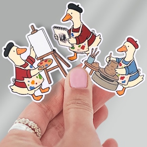 May include: Three stickers featuring cartoon geese dressed as artists. One goose paints on a canvas, another sketches, and the third works on a pottery wheel. Each goose wears a beret and apron with paint splatters, suggesting an art-themed product.