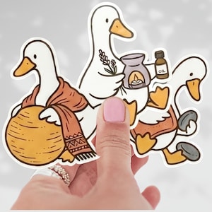 May include: A sticker featuring three cartoon geese. One goose wears a scarf and carries a round object. Another goose holds a diffuser and a bottle labeled "OIL." The third goose carries a small stone. The geese are white with orange beaks and feet.