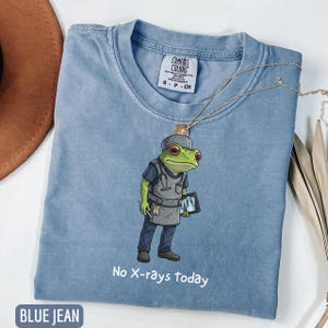 May include: Blue jean Comfort Colors t-shirt featuring a cartoon frog dressed as a doctor, holding a tablet. The shirt has the text "No X-rays today". A gold necklace is draped across the shirt.