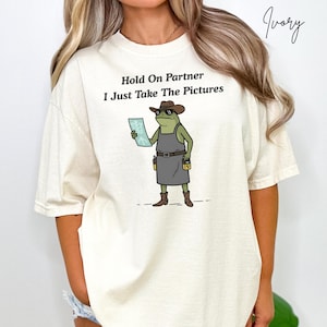 May include: Ivory-colored t-shirt with the text "Hold On Partner I Just Take The Pictures." The shirt features a cartoon frog wearing a cowboy hat, sunglasses, and an apron, holding a document. The word "Ivory" is written in cursive.