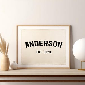 Puede incluir: Un cuadro enmarcado con el nombre "ANDERSON" en letras negras en negrita, con "EST. 2023" debajo. El cuadro tiene un fondo beige y un marco de madera natural. El cuadro está sobre una mesa de madera.