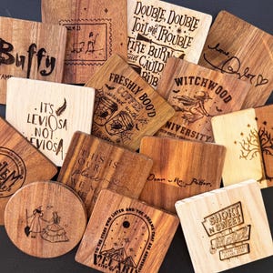 Puede incluir: Una colección de posavasos de madera con varios diseños y textos grabados. Los diseños incluyen frases como "Buffy", "Double, double toil and trouble" y "Short n Sweet Motel". Los posavasos son cuadrados y redondos, con un acabado de veta de madera natural.